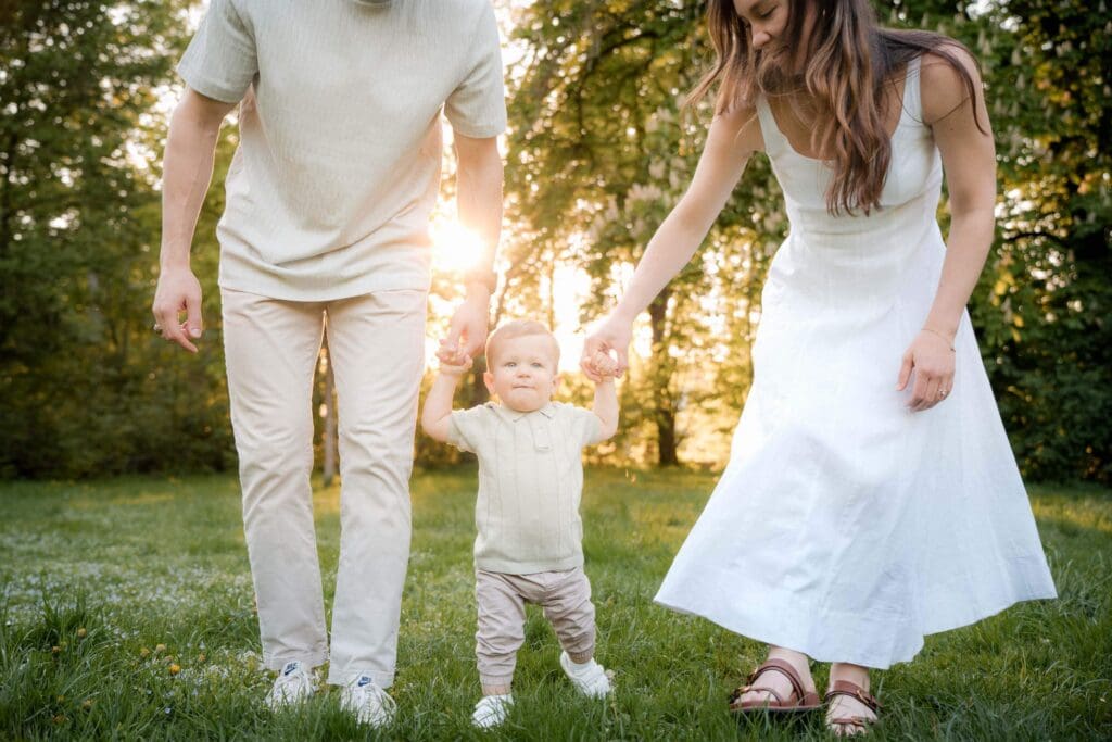 A couple holds hands of one-year-old learning to walk in a sunlit park during golden hour family photoshoot.