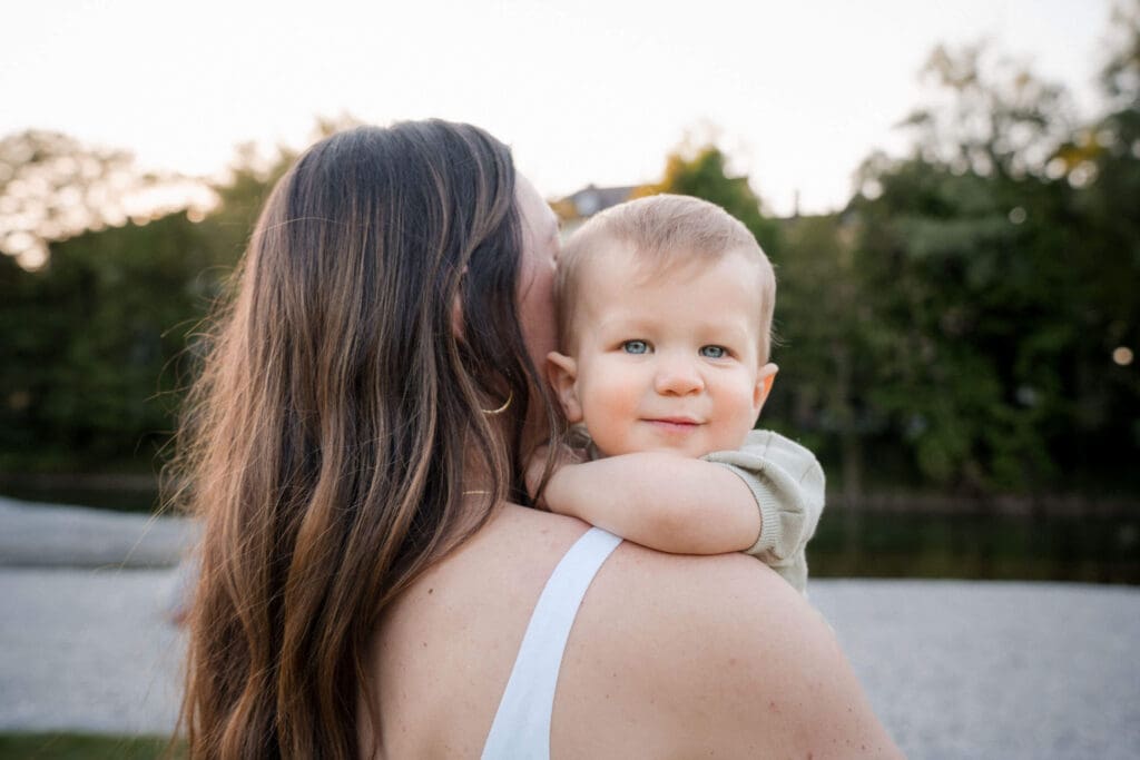 A mom wearing a white dress holds a young child peering over her should, while standing outdoors at sunset near the Isar River.