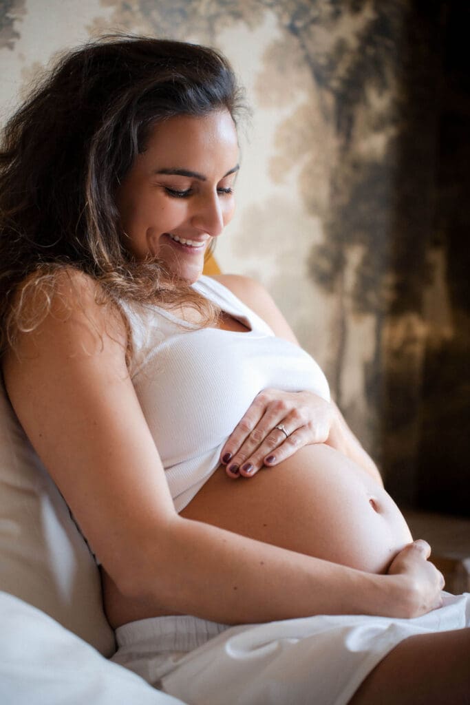 Pregnant woman sits on bed cradling her baby bump while smiling