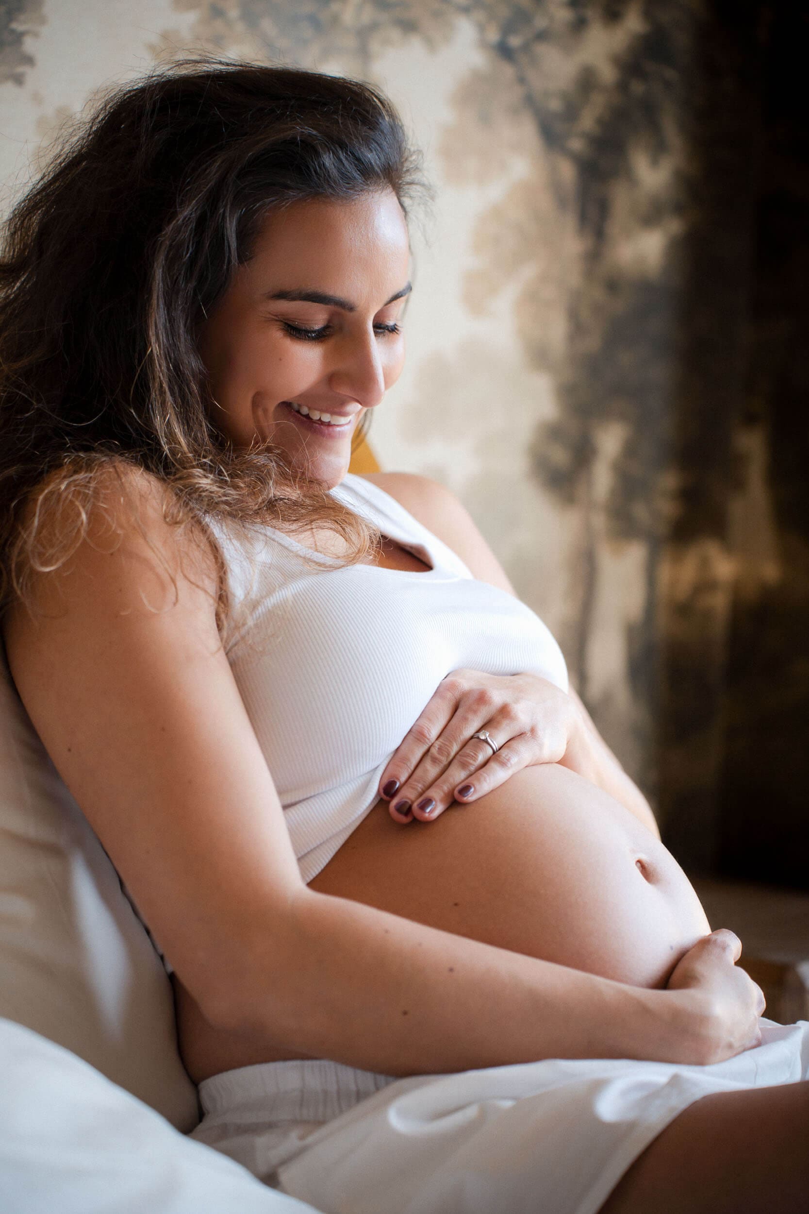 Pregnant woman sits on bed cradling her baby bump while smiling