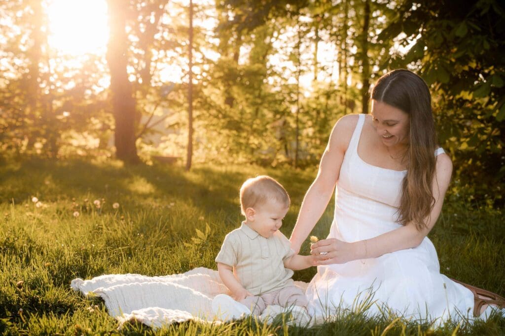 A mom in a white dress sits with a small child in a sunlit meadow during golden hour.