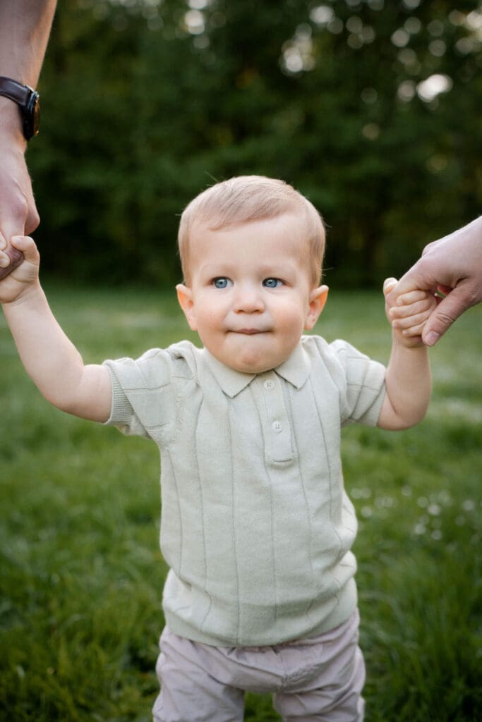Small child in cream knit vest and gray pants holds hands with adults while standing on grass.