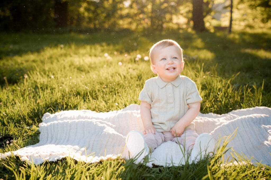 A toddler boy sits on a blanket in a sunlit grassy field during golden hour.