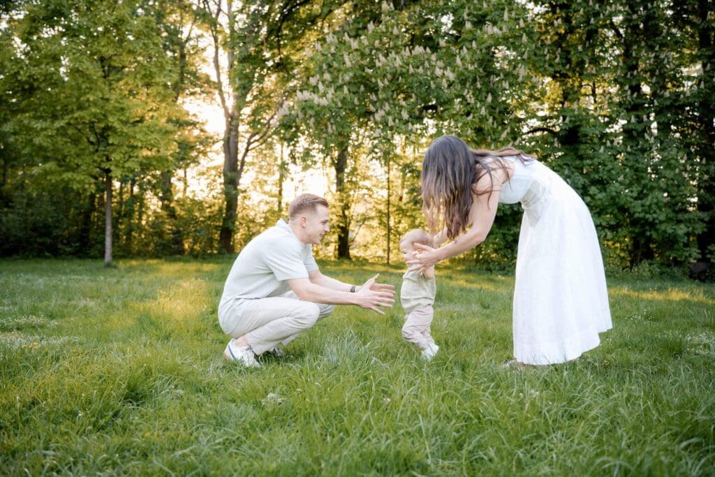 Mom holds one year old's hands as he attemps to walk to dad during summer photoshoot in Munich