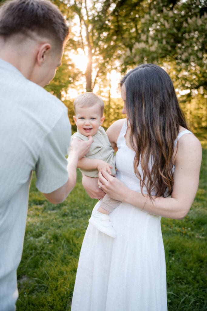 A father and baby in white outfits share a playful moment at sunset in a grassy outdoor setting during a photoshoot.