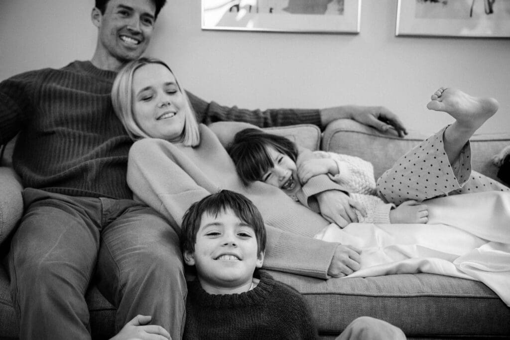 Family cuddles together on couch as little girl laughs sticking out her leg.