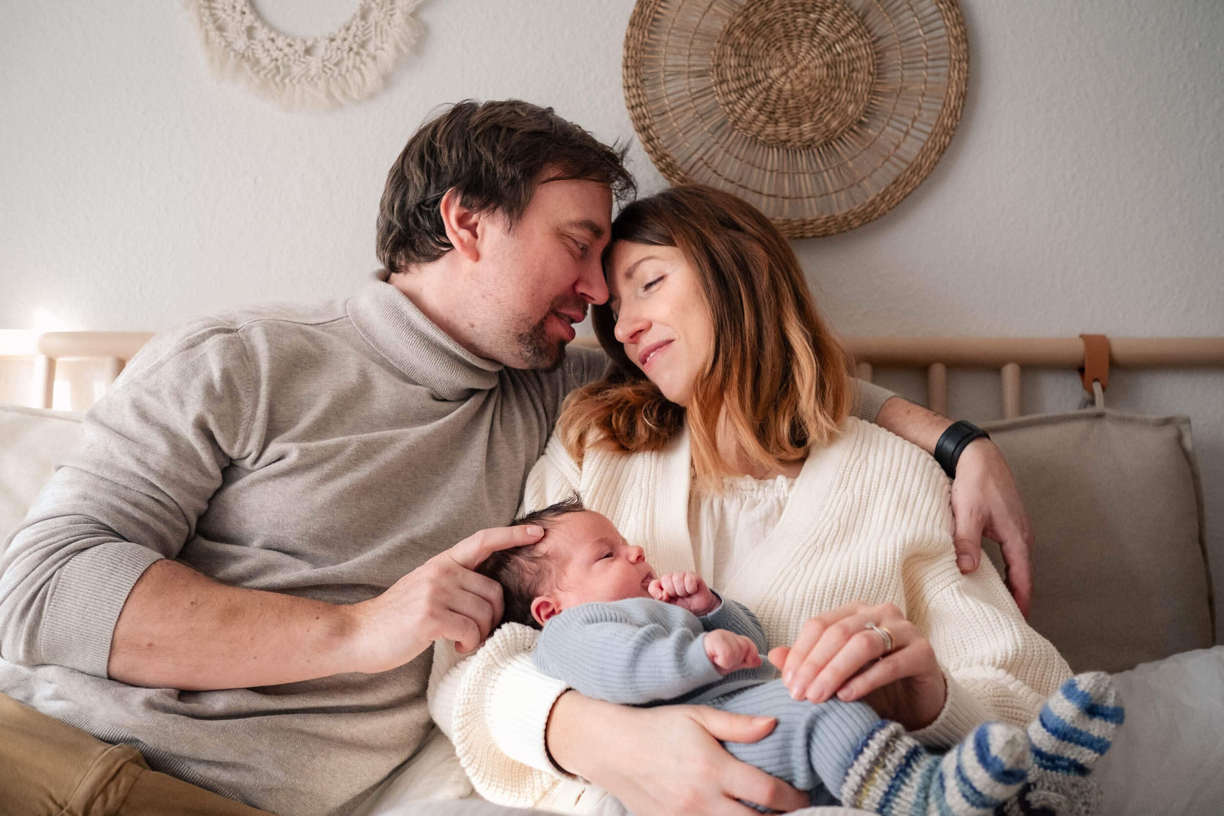 Couple rests on bed while snuggling and holding newborn baby in cozy living room with woven wall decor.