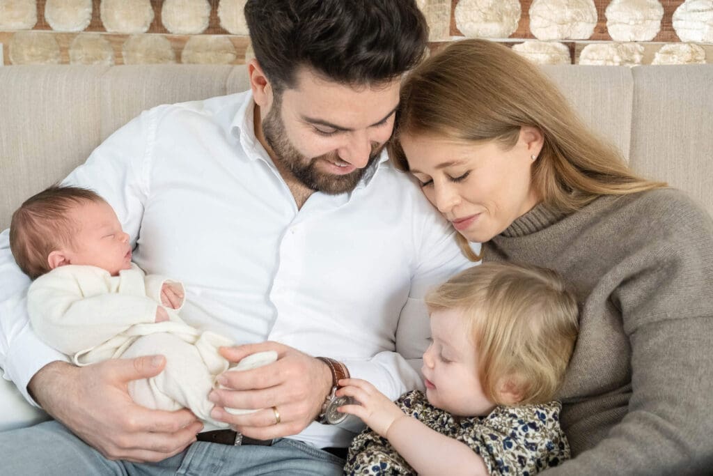 A family rests together on a couch while holding and bonding with their newborn baby and toddler.