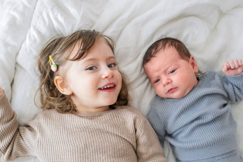 Toddler girl with a strawberry clip in her hair, lays next to her newborn brother in their parents bed.