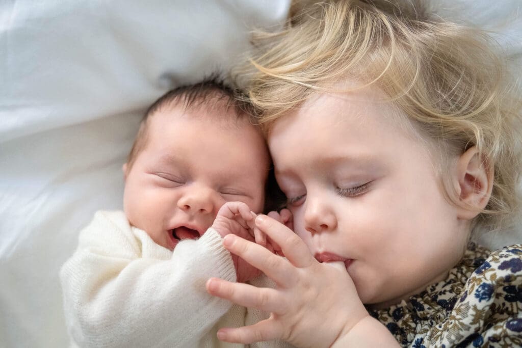 Toddler girl lays next to newborn baby and closes her eyes while sucking on her thumb.