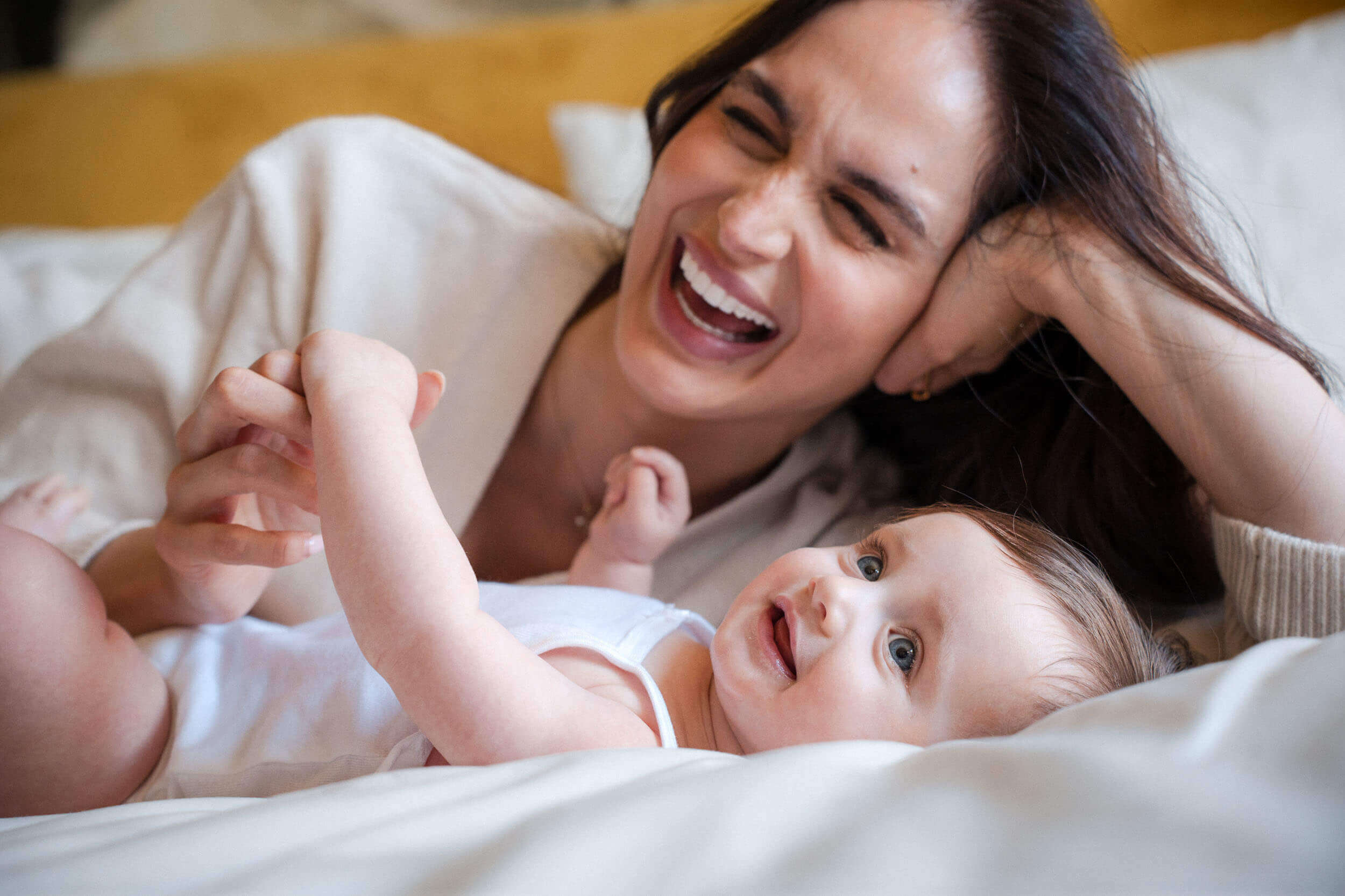 Mother and baby lying on white bedding share joyful laughter while playing together.