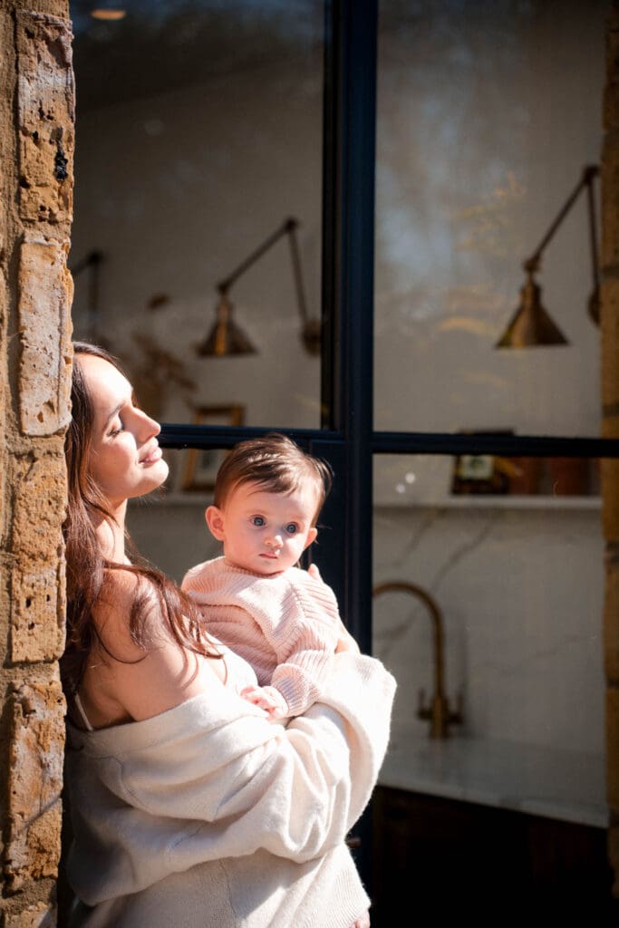 Mother holds baby while leaning back against exposed brick wall soaking in sunshine on her face.