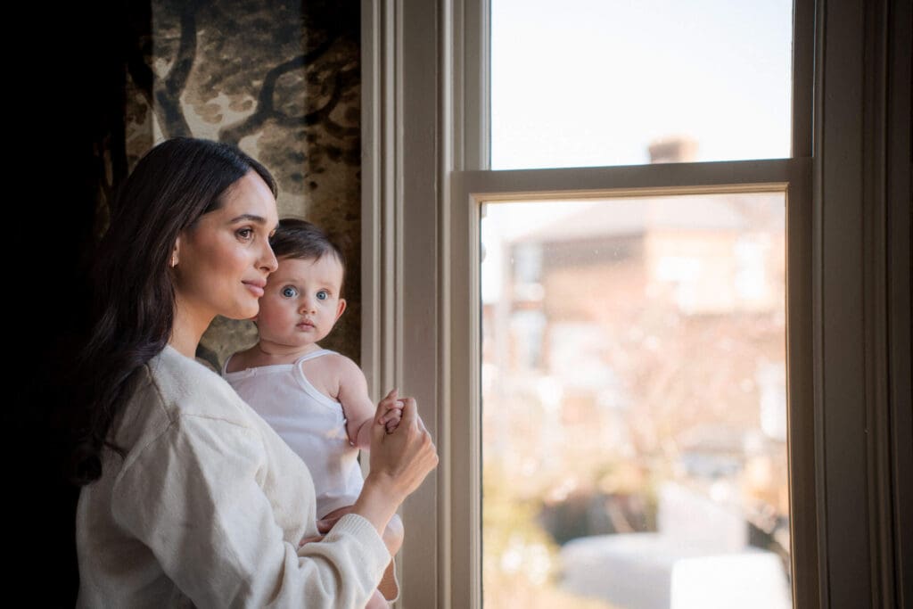 A mom in cream sweater holds a small baby while gazing out a large window with natural light streaming in.