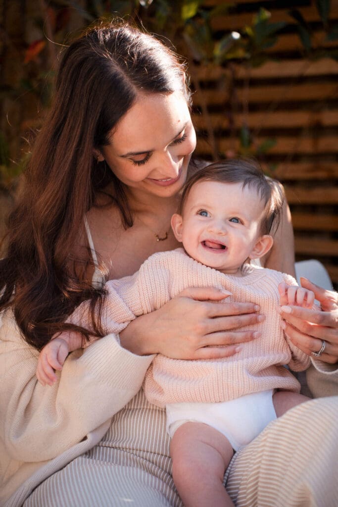 Mother and smiling baby in matching light colored sweaters share a tender moment on a sunny day.