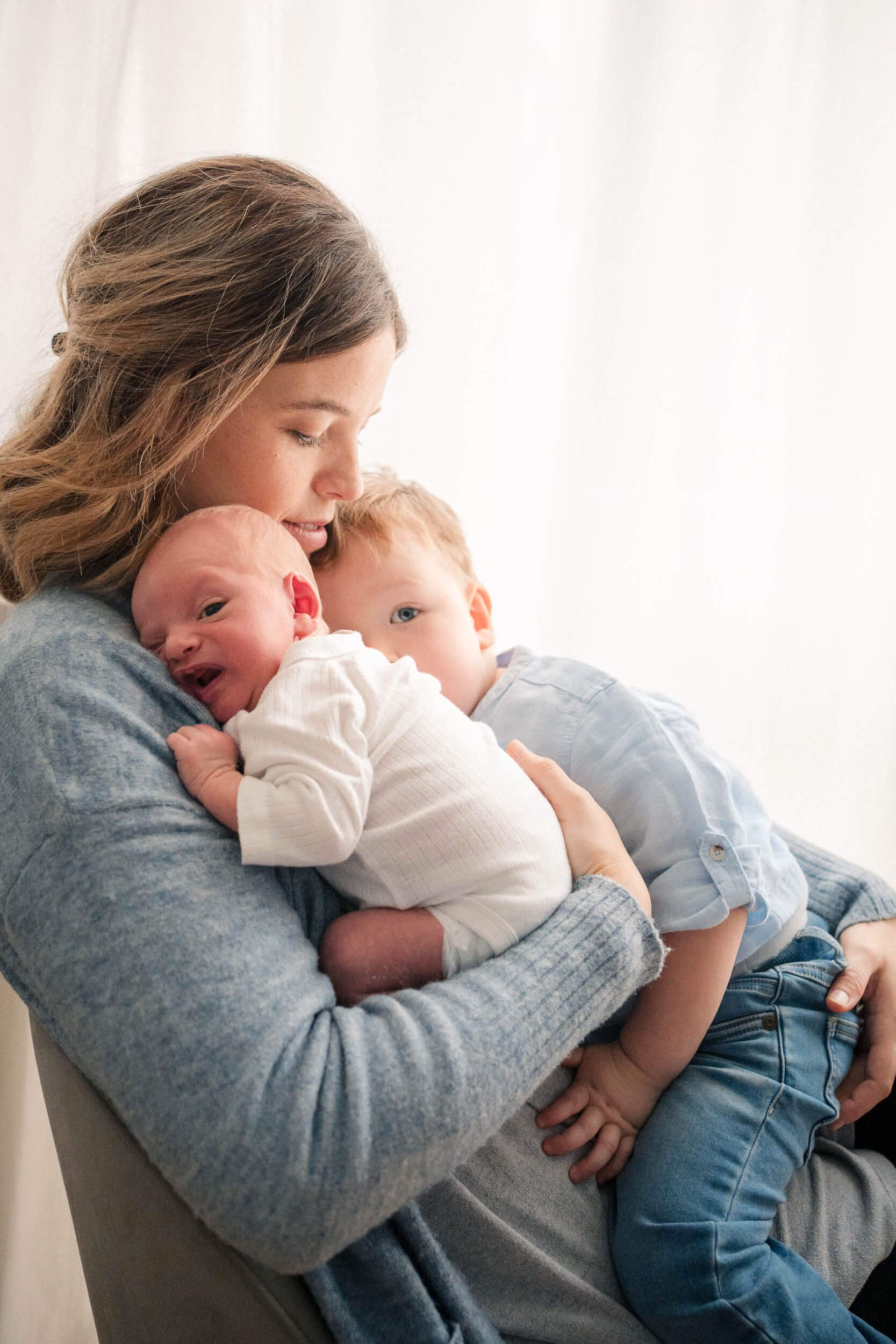 Mama kuschelt ihr Baby gleichzeitig mit ihren Kleinkind auf ihren Schoß gegen ein sehr helles Fensterlicht