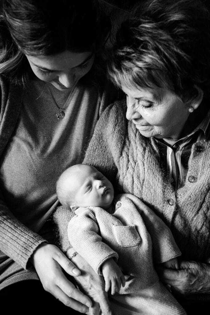 Schwarzweißporträt dreier Generationen, die einen intimen Moment beim Anblick eines schlafenden Babys teilen.