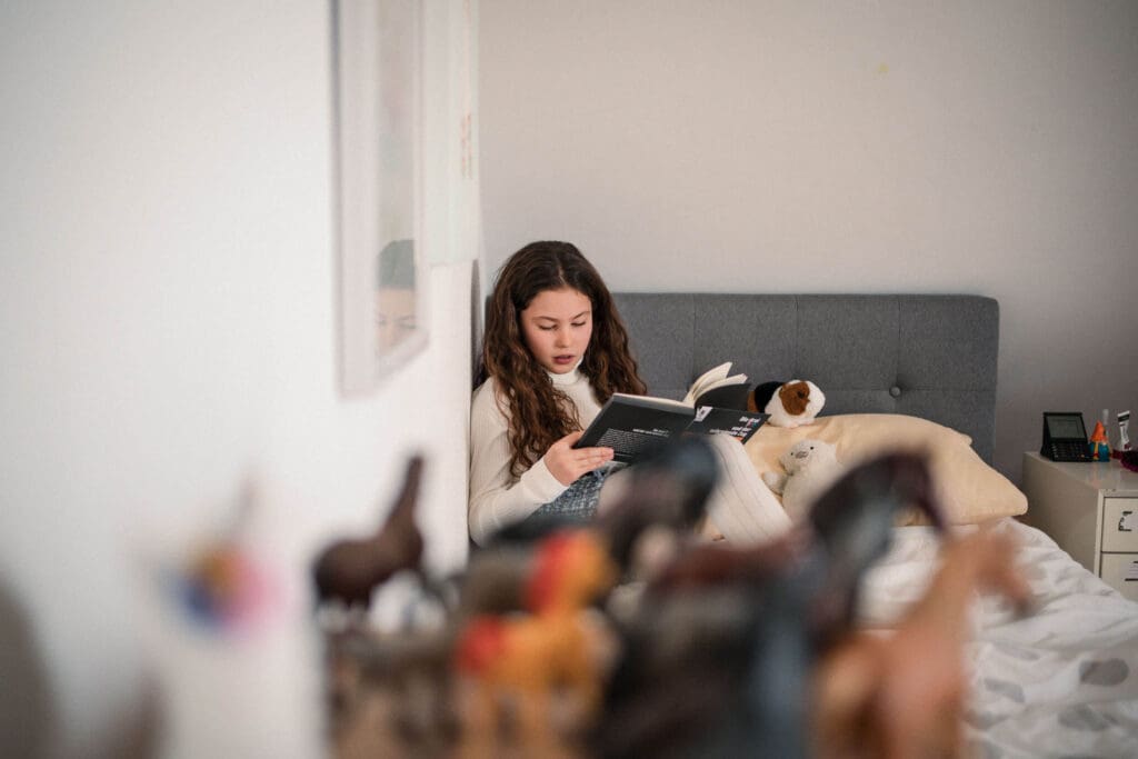 A girl relaxing in bed reading a book while surrounded by plush toys and cushions in a cozy bedroom setting.