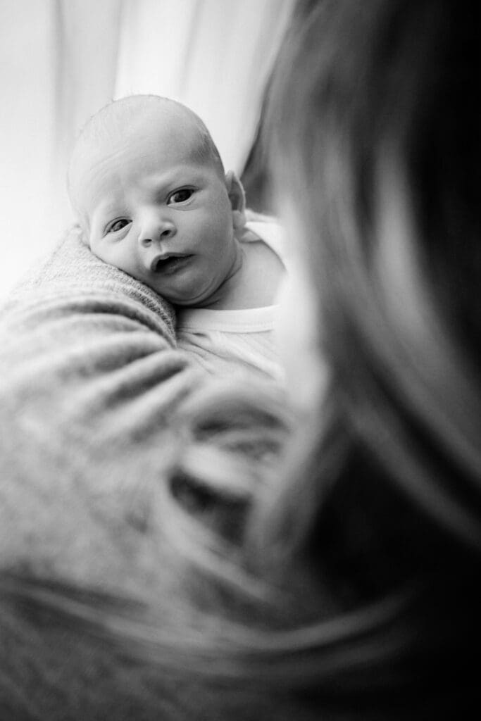 Schwarz-Weiß-Porträt eines Newborns, der in den Armen der Mutter gehalten wird und verwundert zu ihr aufblickt