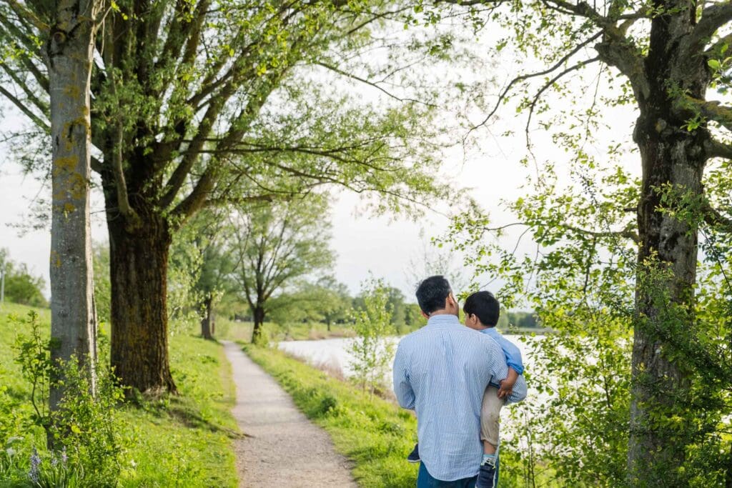 Father carries young son on hip as he walks along path next to Riemersee in East Munich.