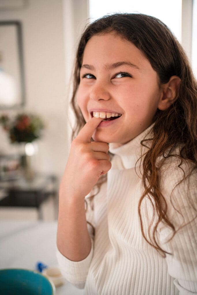 A young girl in a white sweater smiles while touching their chin in a bright indoor setting.