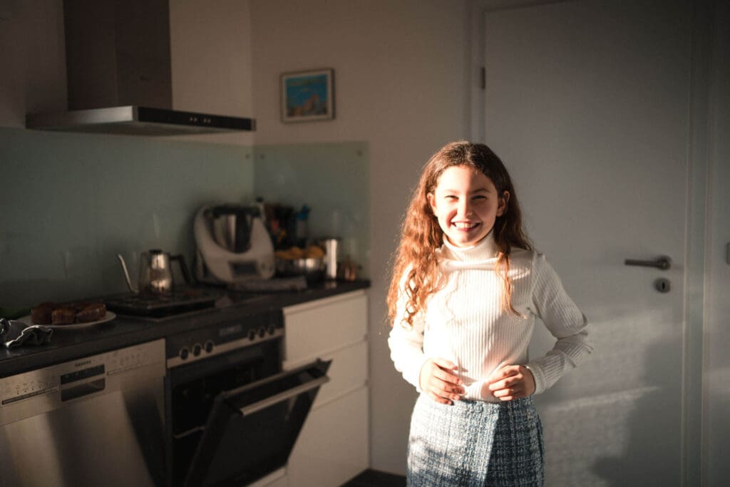Girl in a bright kitchen wearing a white sweater and patterned skirt standing by a modern stove and range hood.