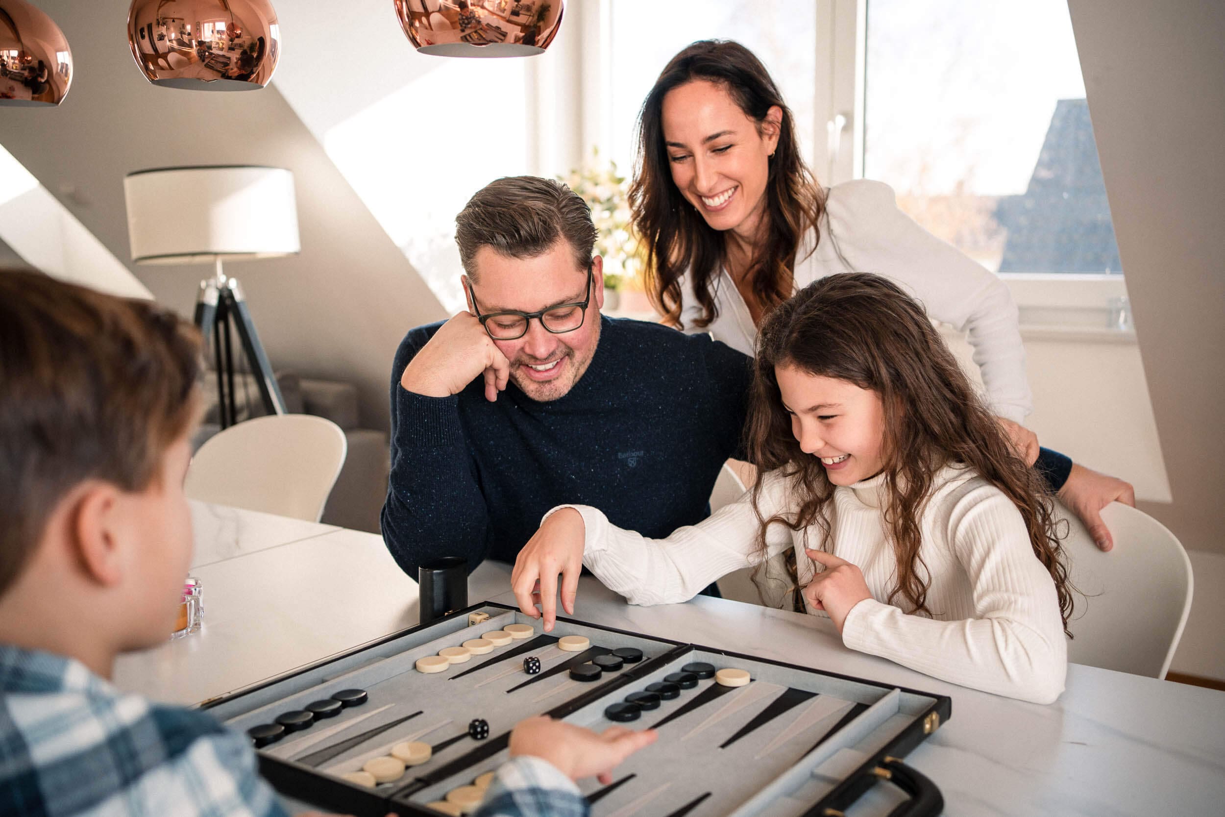 A family gathers around a table playing a competitive game of backgammon together in a bright living room.