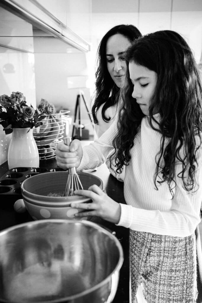 A black and white photo showing a daughter and mom whisking ingredients in bowls together in a bright kitchen.