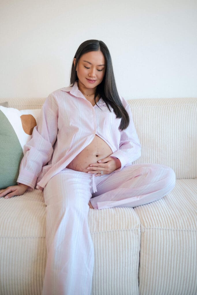 Pregnant woman in pink pajama set sits comfortably on cream-colored couch cradling her baby bump.