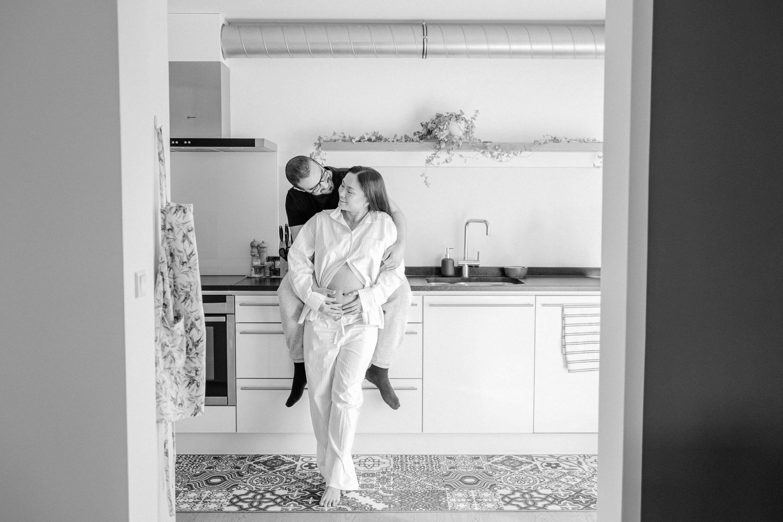 Black and white photo of a couple embracing in a modern minimalist kitchen interior, while the woman cradles her pregnant belly.