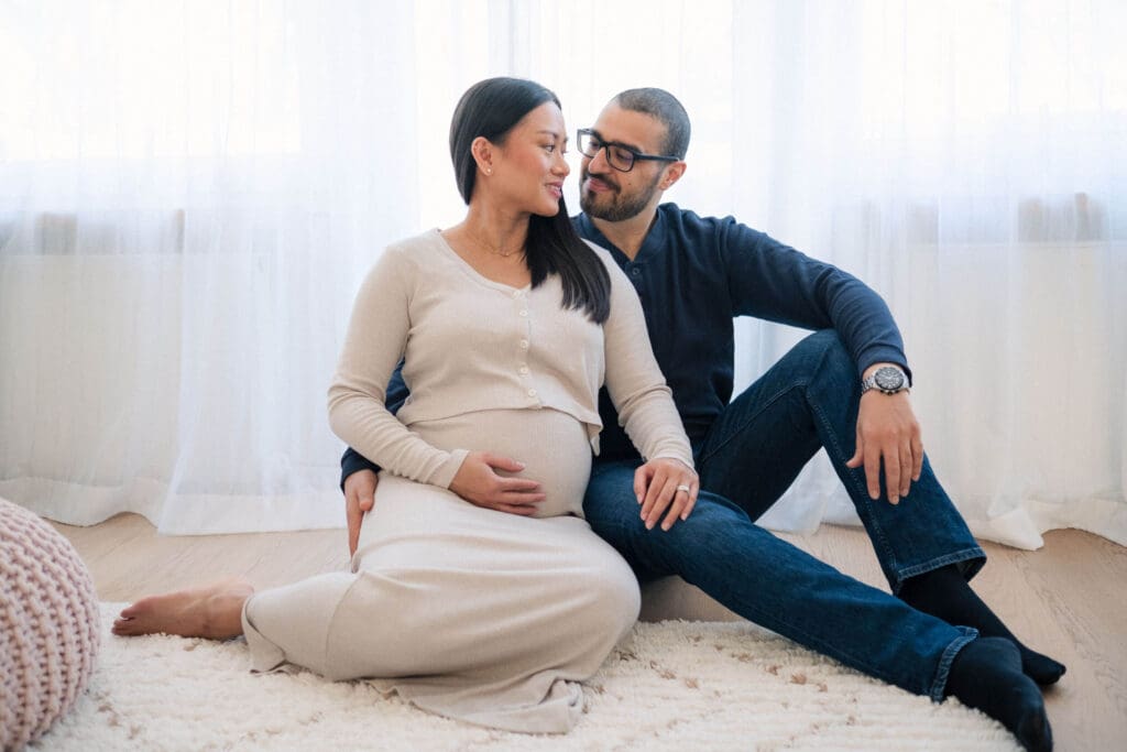 Expecting couple sits together on floor at home during maternity photoshoot, intimate indoor setting with natural light.