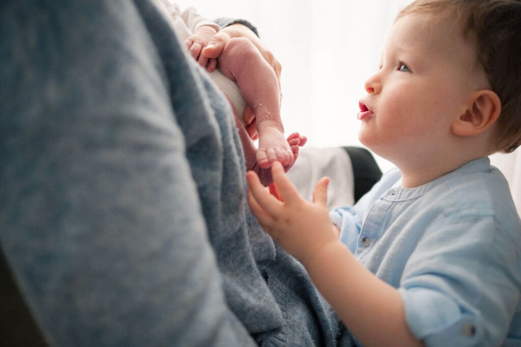 Ein kleines Kind in hellblauer Kleidung streckt seine kleinen Hände nach den Füßen eines Neugeborenen aus