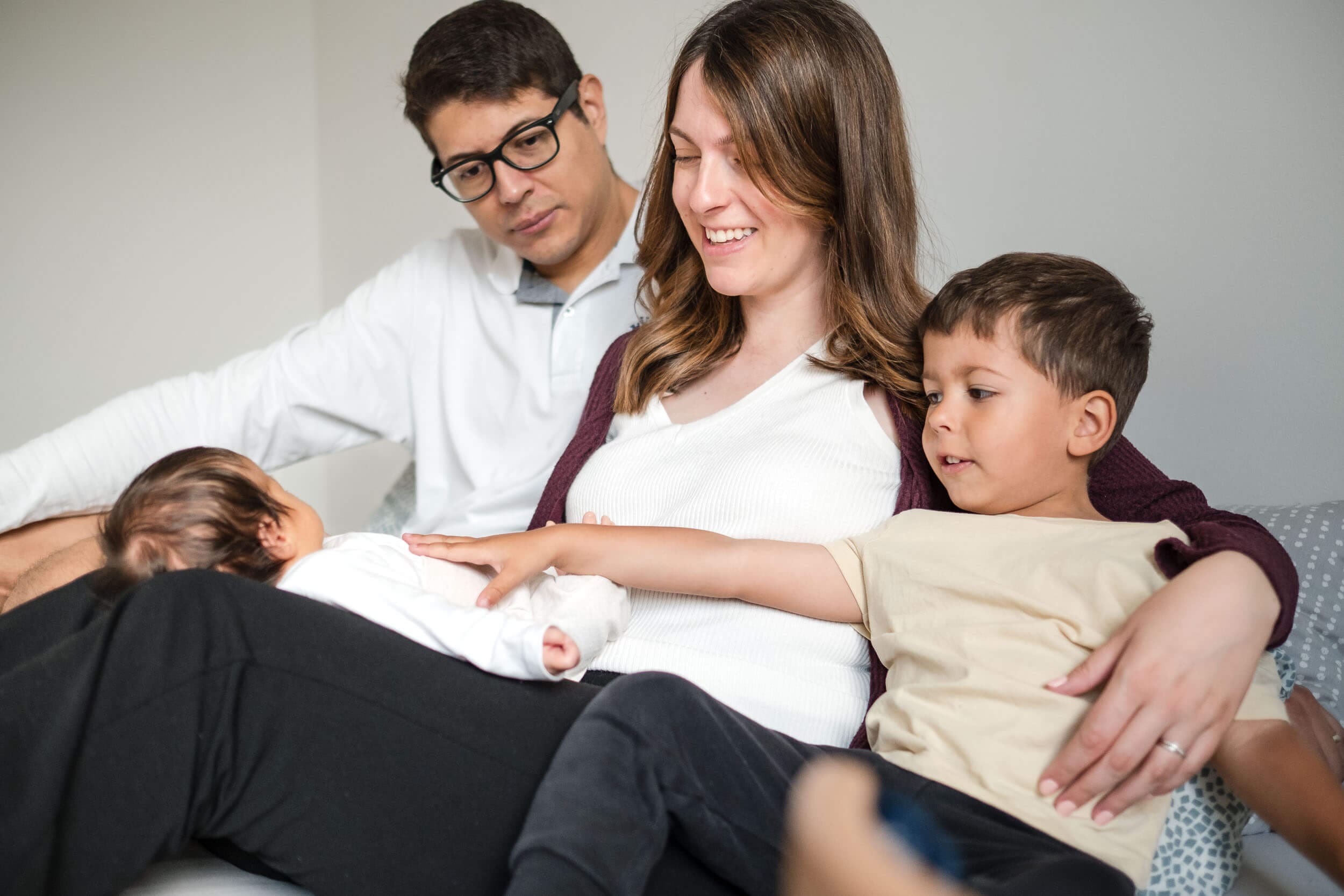 Newborn baby on mother’s lap as family cuddles on bed during in-home newborn session