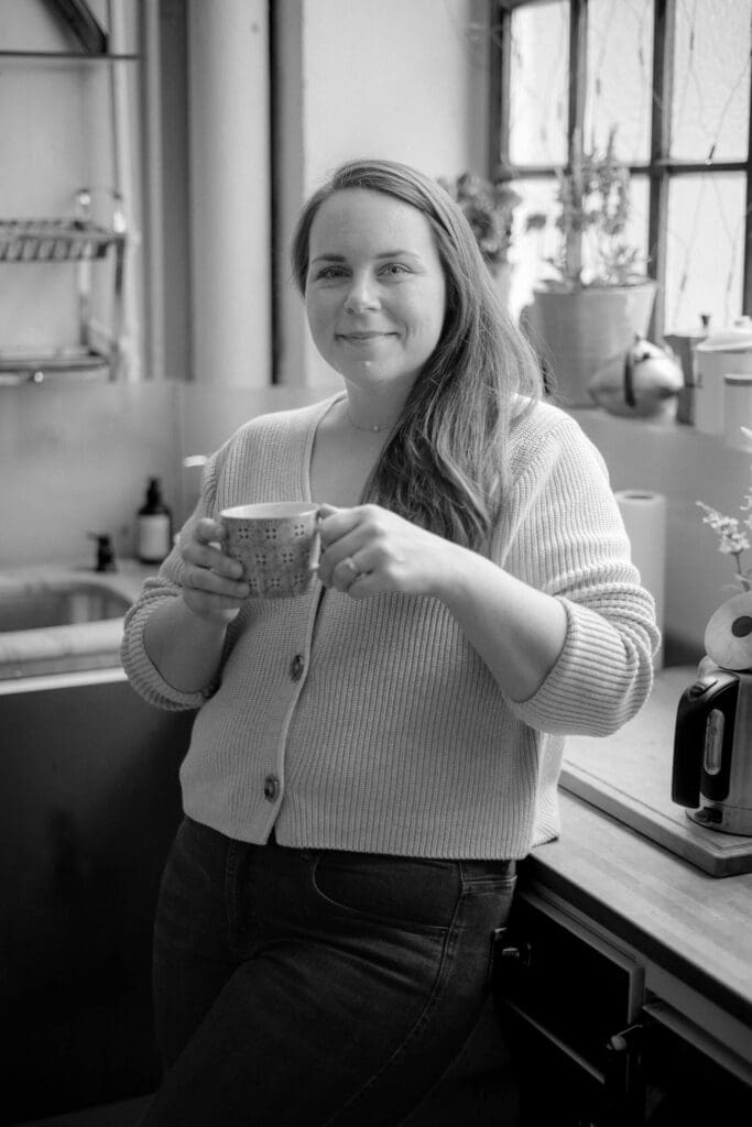 A woman stands in a warmly lit kitchen, holding a coffee mug and smiling gently at the camera. A relaxed portrait of a family photographer in her everyday space.
