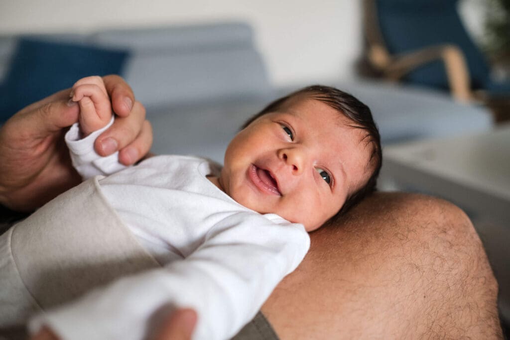 Newborn baby lying on dad's lap, holding his hands and smiling while gazing towards the light from the window.
