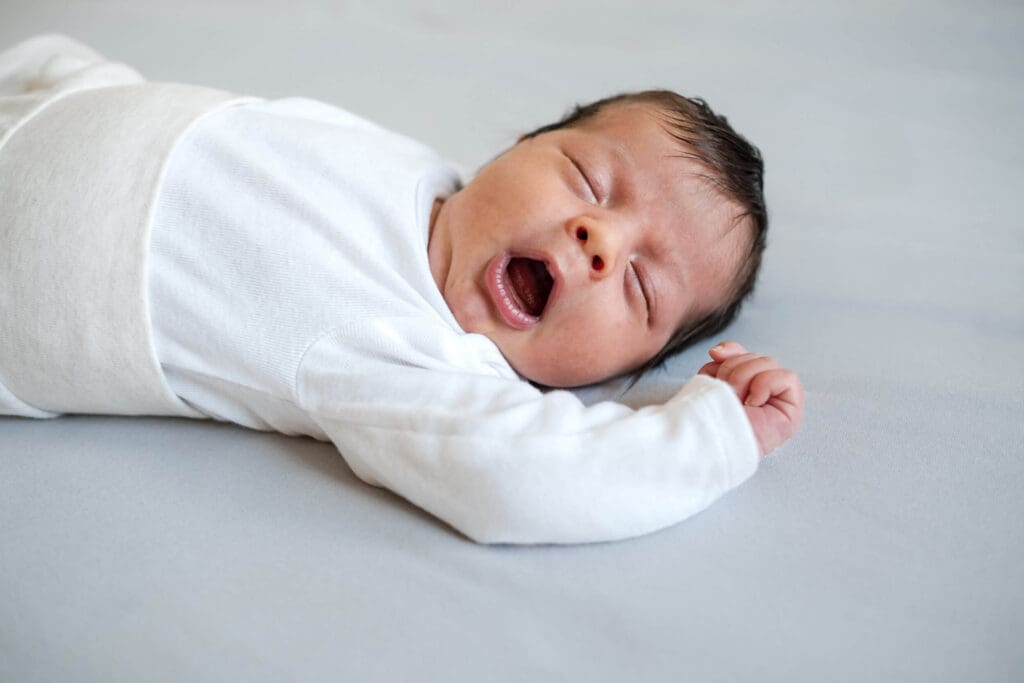 Newborn baby yawning and stretching on bed in a simple white outfit.