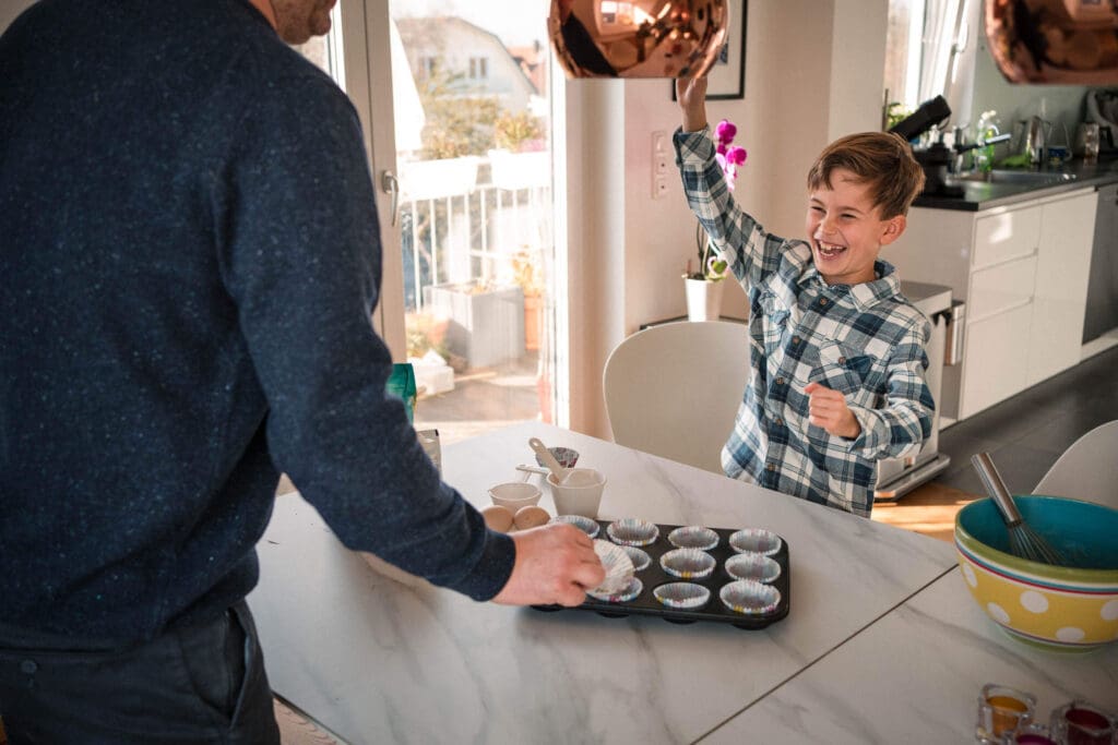 A child excitedly raises a frosting tip while baking cupcakes with an adult in a bright kitchen.