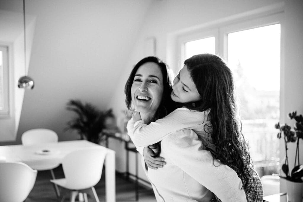 Black and white photo of two people embracing and laughing together in a bright modern room with white chairs.