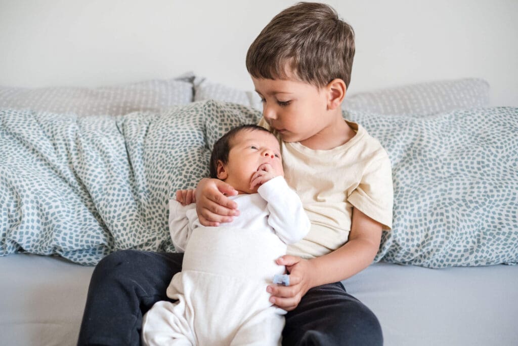 Toddler brother holds newborn sister on his lap for the first time, gently gazing at her face.