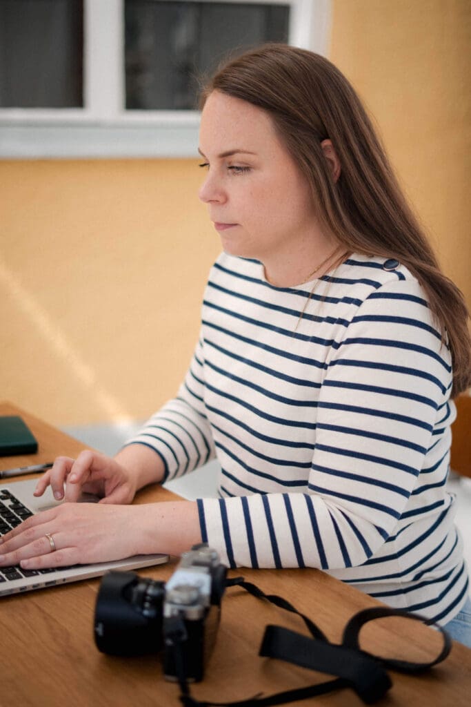 Munich photographer in striped shirt sits at a desk working on a laptop with a retro camera in front of yellow wall