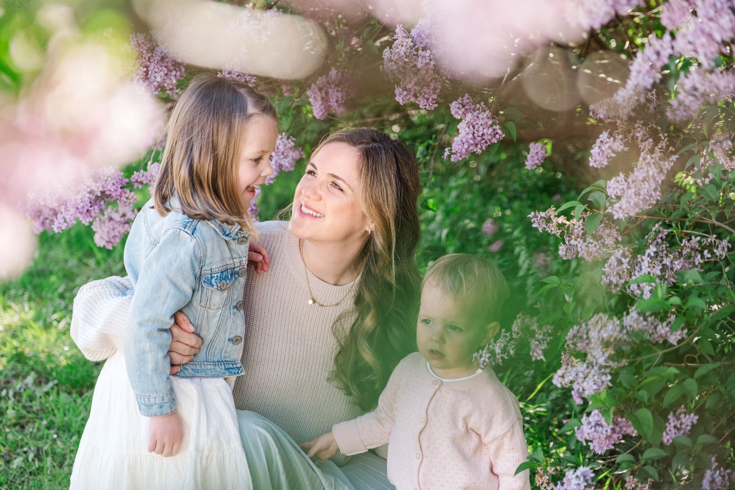 Mom looks lovingly at daughters while kneeling next to Lilac bushes outside of Nymphenburg Palace