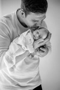 Ein zartes Schwarz-Weiß-Porträt eines Elternteils, das ein in weiße Kleidung gewickeltes Neugeborenes sanft wiegt und küsst.