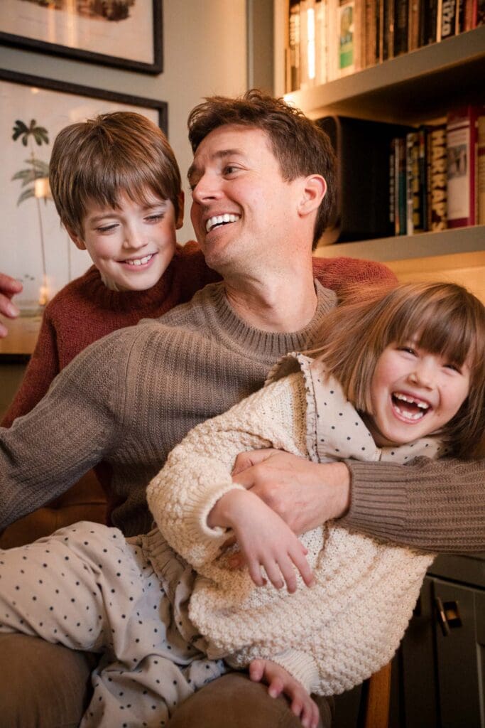 Kids laughing piled on top of their dad in coordinating muted color sweaters