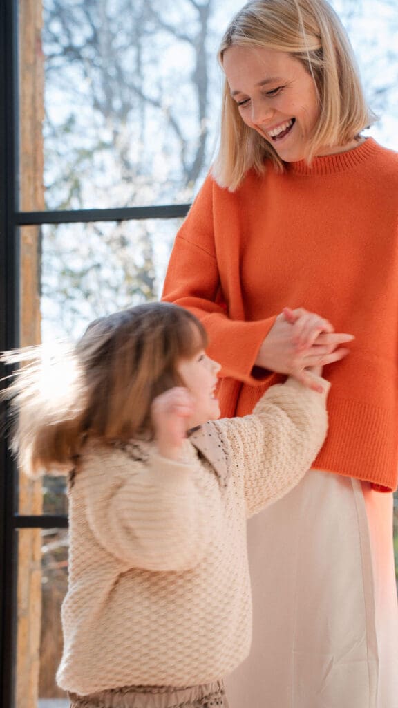 Mother in orange sweater spings daughter while dancing during an in-home family photoshoot