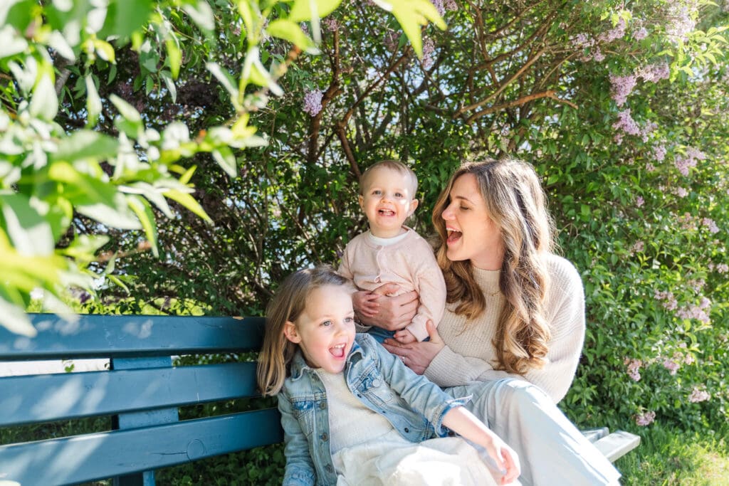 Mom sitting on a bench in from of lilac bushes with two young daughters laughing while they have a tickle fight