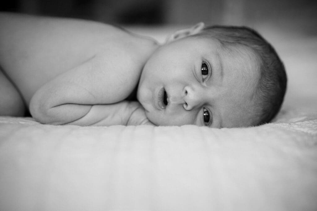 Schwarz-Weiß-Porträt, das ein kleines Baby zeigt, das auf einem Tuch liegt und mit strahlenden Augen direkt in die Kamera blickt.