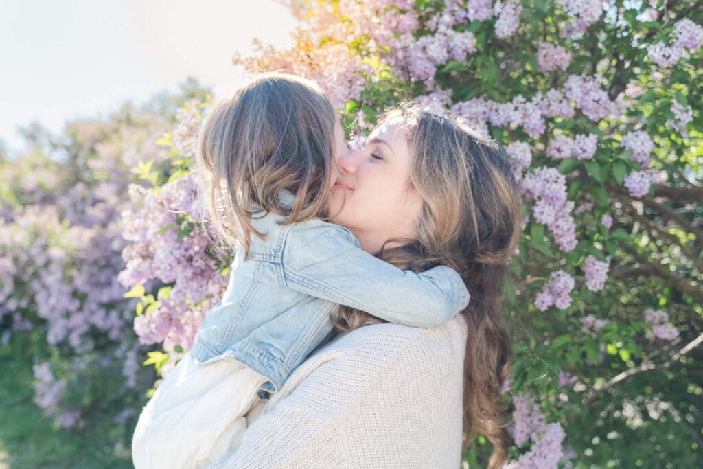Mom standings in front of a lilac bush with the sun peeking behind while she squeezes her daughter tightly,
