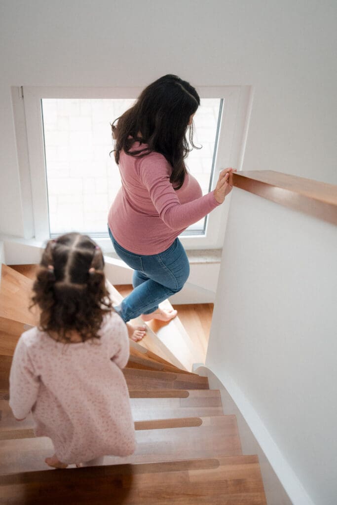 Pregnant mother climbing wooden stairs at home with young daughter following close behind during a lifestyle maternity session in Munich