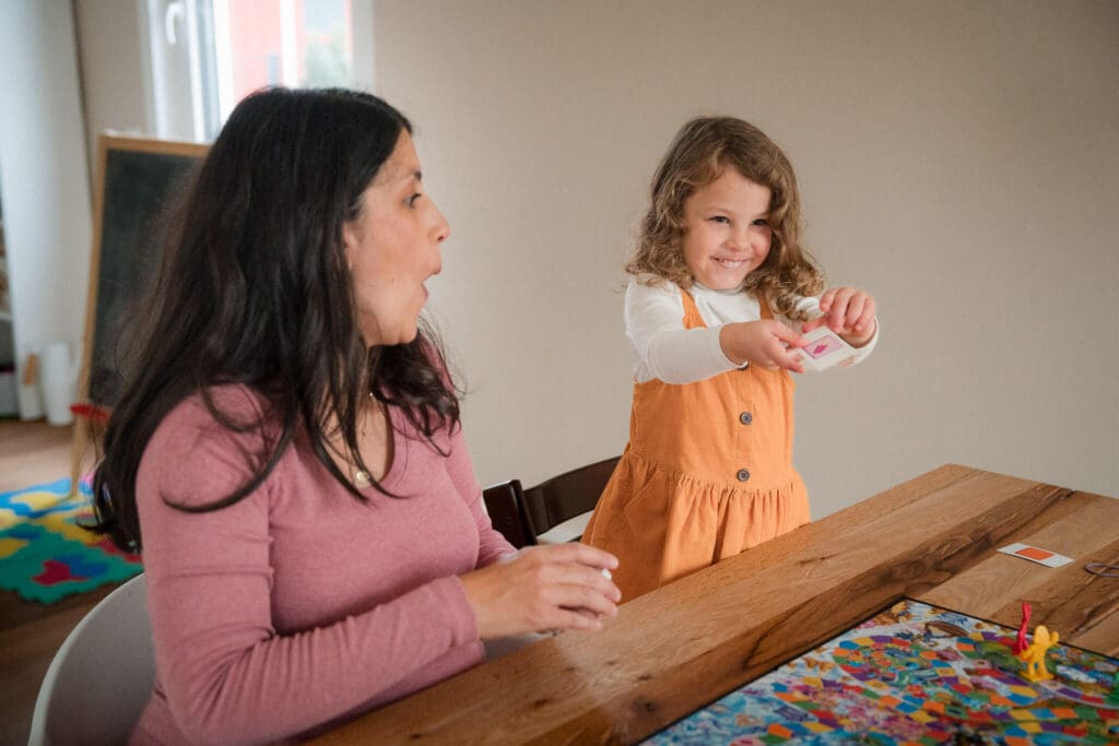 Mother laughing while her young daughter proudly shows her a board game card at the kitchen table during an in-home family session in Munich