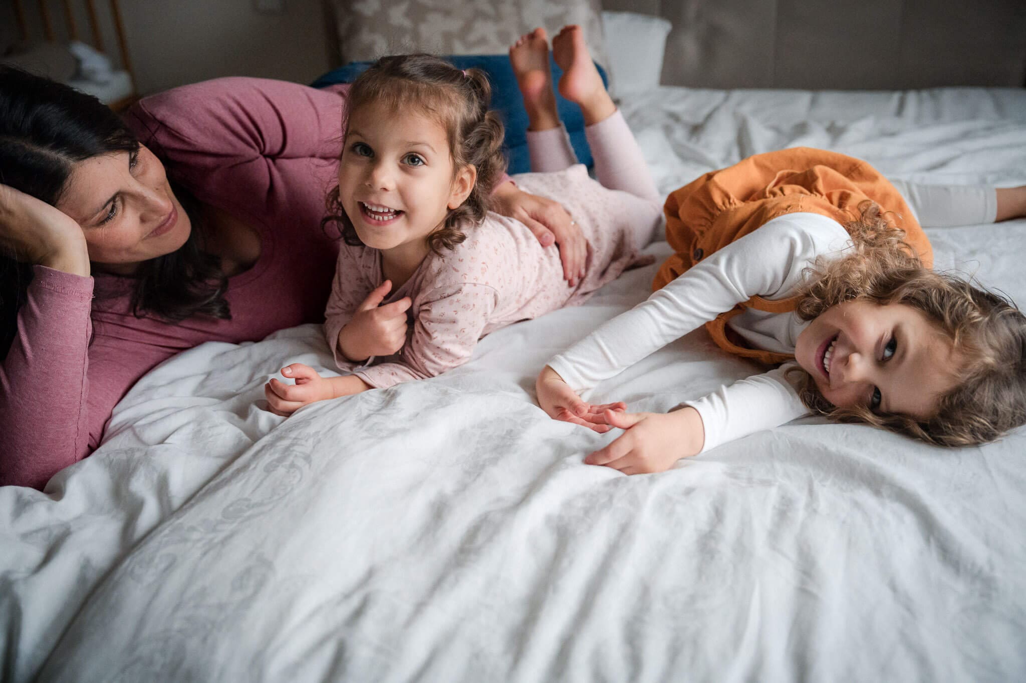 Mother lying on a bed laughing as her two young daughters play and tumble around her during an in-home family photoshoot in Munich