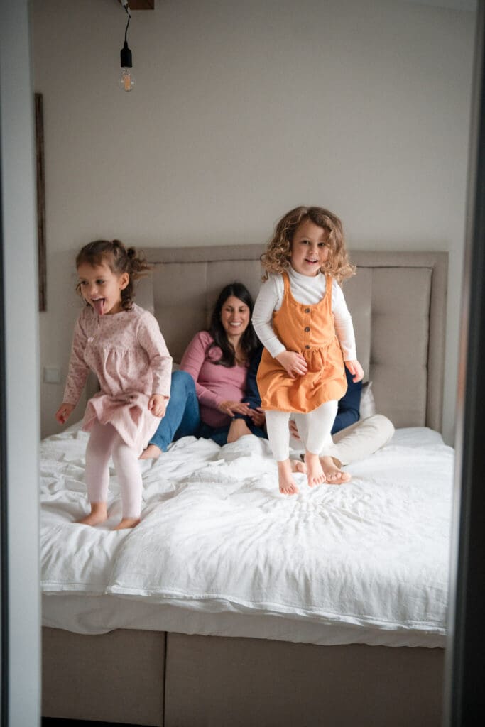 Two young girls jumping on the bed laughing while parents watch and smile during an in-home family photoshoot in Munich
